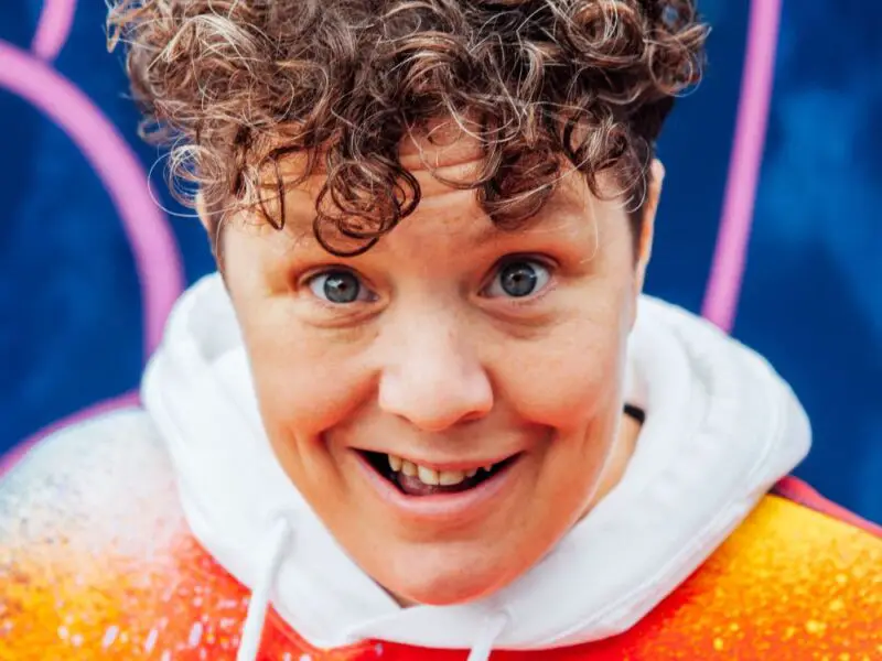 A close up of Jess Thom, who has short, curly, brown hair, wearing an orange jacket over a white hoodie standing in front of a blue and pick background pulling a startled face