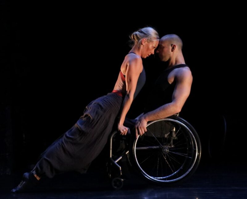 Joel Brown and Eve Mutso dancing on a black stage with Joel in a manual wheelchair wearing a black vest and Eve balanced on his lap facing him wearing a black sarong and with her blonde hair tied up