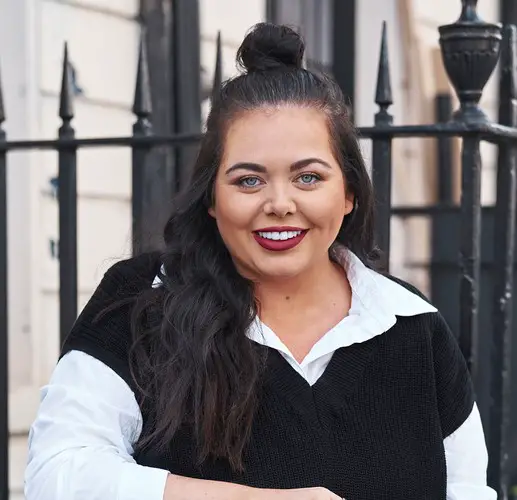 Scarlett Moffatt standing in front of black iron railings wearing a white shirt with a black jumper over the top and with half of her long black hair tied in a top knot and the rest across her left shoulder