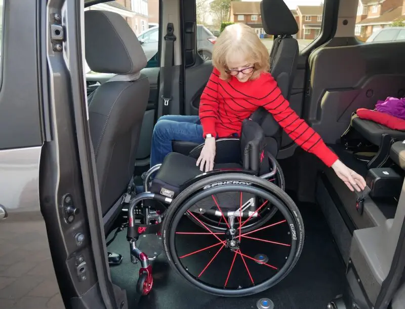 Susan with shoulder length blonde hair wearing blue jeans and a red top, sat in the rear passenger seat of our wheelchair accessible vehicle next to her manual wheelchair