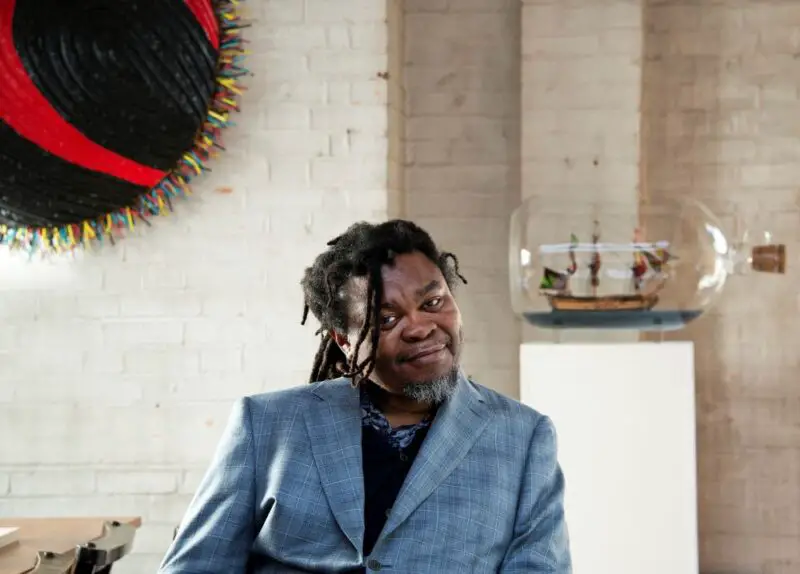Yinka Shonibare, who is black and has dreadlocks, wearing a grey suit jacket and a black top, sat in a modern room with art on the wall behind him