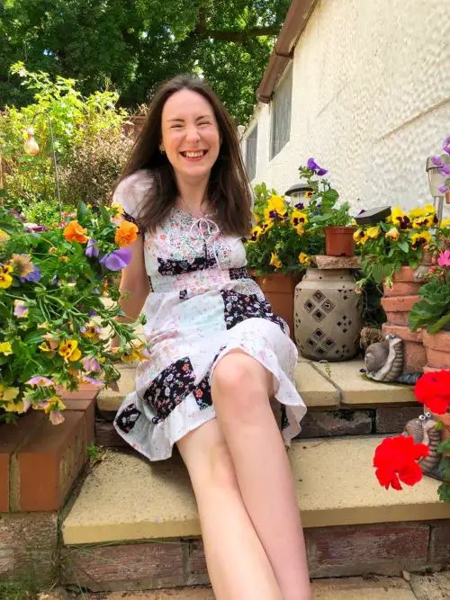 Rebecca Sullivan in a white and black patchwork dress with her long, brown hair down, sat on steps in her garden surrounded by plants in pots