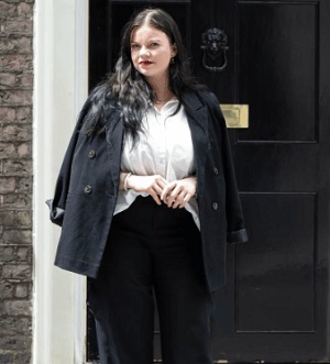 Verity with long black hair wearing a white blouse, black jacket and black trousers standing in front of No 10 Downing Street