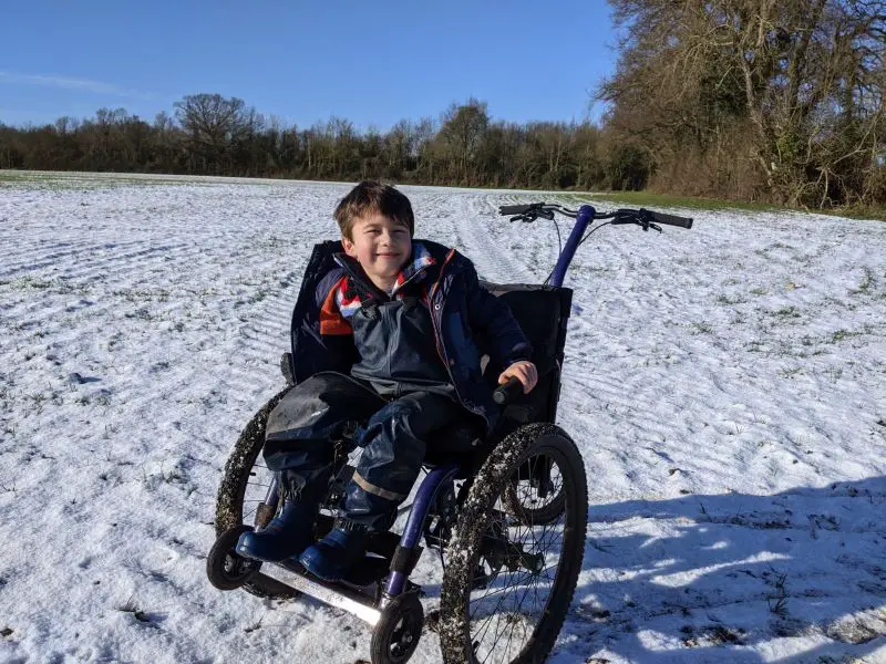 A young boy with dark hair wearing a black coat and waterproof trousers sat in a Mountain Trike all-terrain wheelchair in a snow covered field