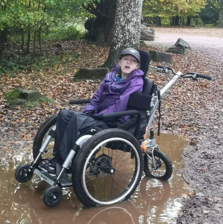 A young girl wearing a cap, purple coat and waterproof trousers sat in a Mountain Trike all-terrain wheelchair in the woods