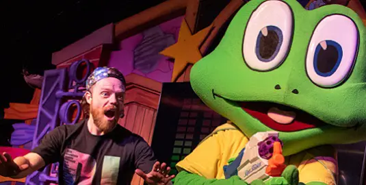 A man with red hair and a beard wearing a black T-shirt and bandana looking surprised as Freddo the frog - a giant green frog -points a water gun at the camera