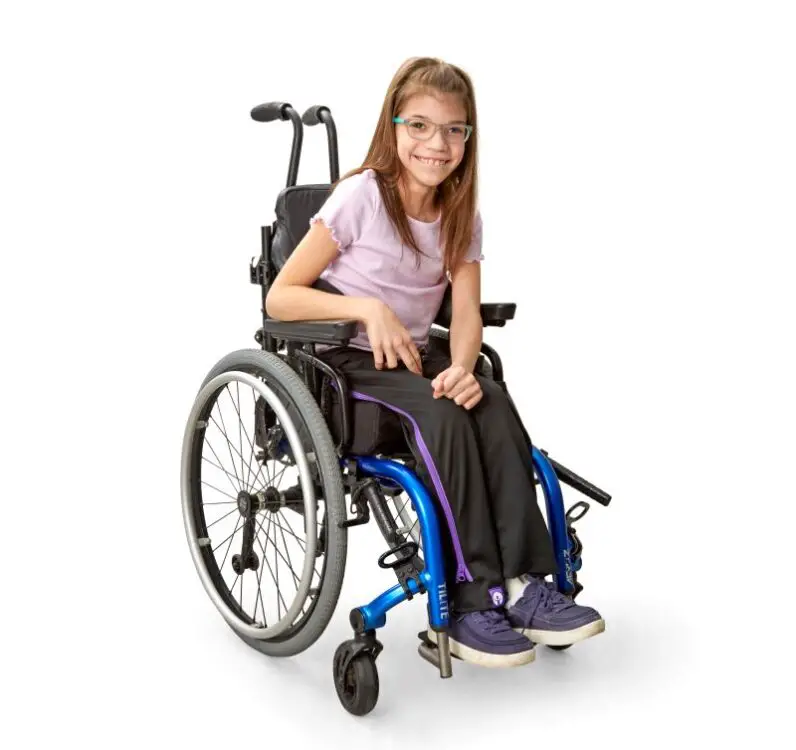 Stella with long brown hair in her wheelchair smiling. She is wearing a pale pink T-shirt, ZipOn adaptive trousers, pale blue glasses and navy blue shoes. 