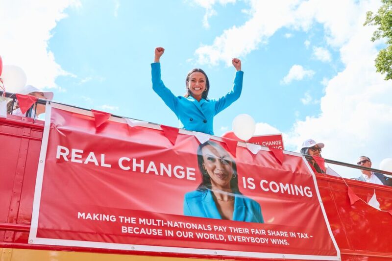 Natalie standing on an open top London bus campaigning with her arms up smiling. There is a poster on the bus with an image of Natalie and text reads, real change is coming making the multi nationalist pay fair share tax because in our world, everybody wins