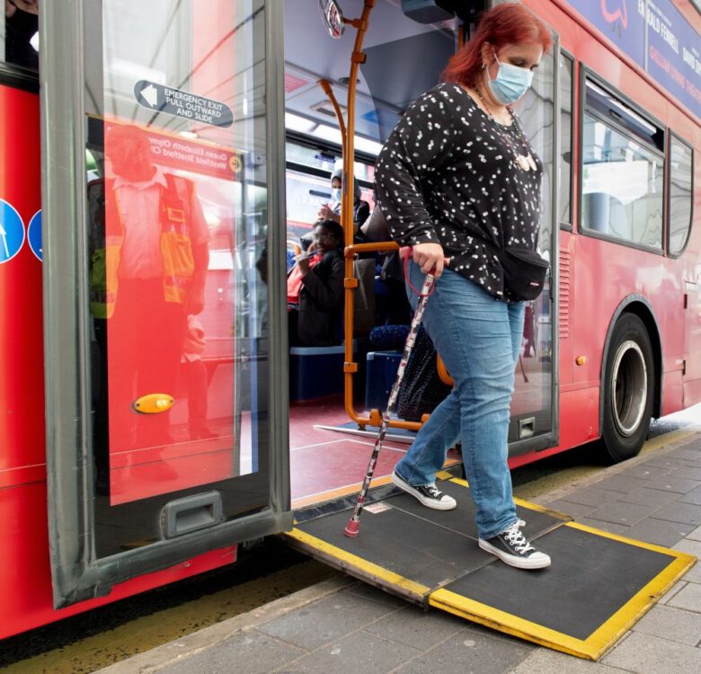A woman with red hair wearing a black top and blue jeans using a stick-getting off a bus in London using a ramp