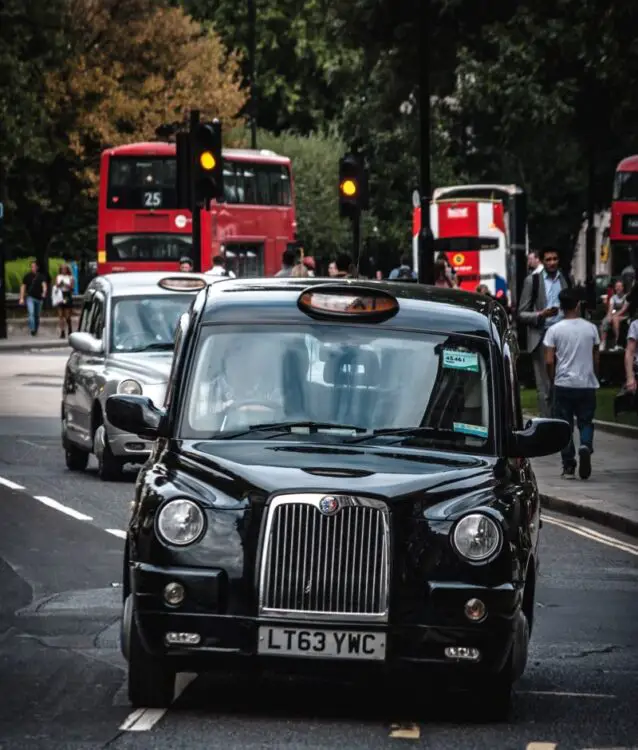 A black taxi in London with red busses behind it