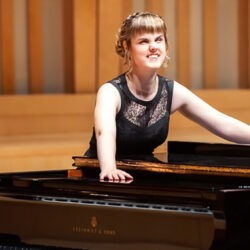 Rachel Starrit in evening attire, leaning casually on a grand piano on stage
