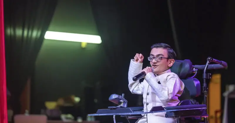 Image is a photograph of Sparsh Shah on stage with microphone in hand