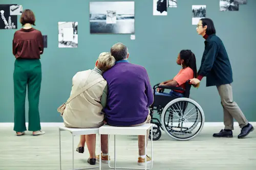 Couple sitting on a bench admiring art work. In the background is a person pushing someone in a wheelchair
